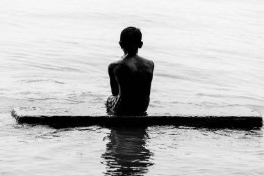 Bir çocuk banyo yaparken nehir suyunda bir kayanın üzerinde oturuyor. Güneş ışığına karşı görüntü. Siluet resmi.