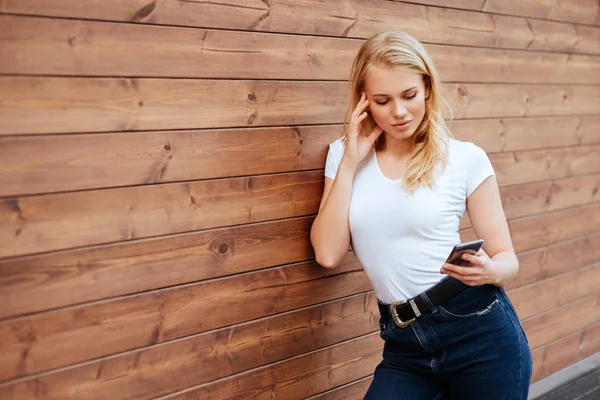 Çekici ahşap duvar arka plan ile kopyalama alanı alanı, cep telefonu ile gülümseyen ve sevinç içinde çok mutlu hissediyorum seyir hipster oldukça raslantı giyimli kadın ayakta kadın gülüyor