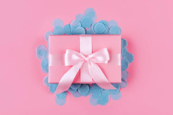 Present box with pink bow on pastel pink background with multicolored confetti. Flat lay style.