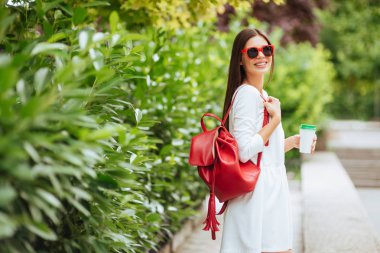 Sokakta yürüyen güzel esmer kız portresi. Bir yandan ve kırmızı çanta başka bir paket servisi olan restoran içki tutmak. Gülümseyerek. Kentsel şehir sahne. Sıcak güneşli havalarda. Açık havada