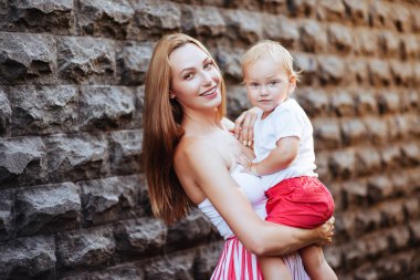 Güzel bayan ve onun tatlı küçük bebek gülümseyerek, önce gri duvarın ayakta portresi. Bebek kameraya bakıyor