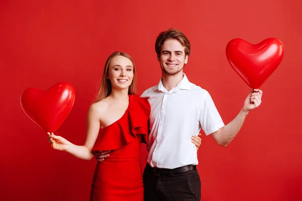Retrato de atractiva pareja joven posando sobre fondo rojo y sosteniendo globos corazón . — Foto de Stock