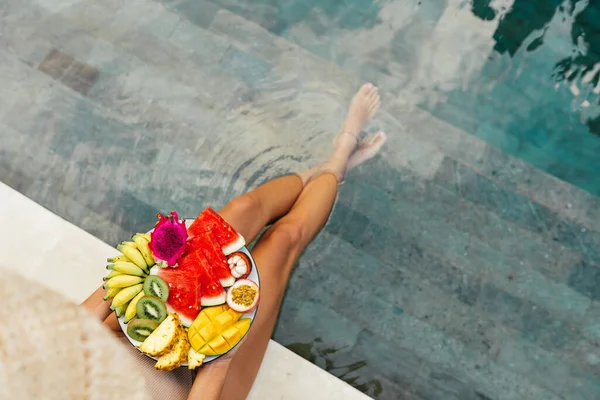 Menina relaxante e comendo prato de frutas na piscina do hotel. Dieta de verão exótica. — Fotografia de Stock