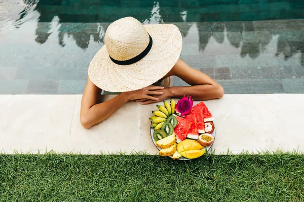 Menina relaxante e comendo prato de frutas na piscina do hotel. Dieta de verão exótica. — Fotografia de Stock