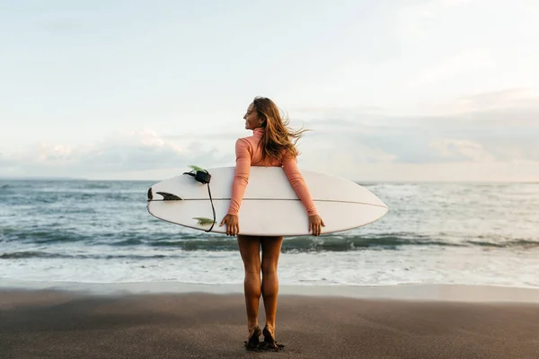 Ung surfare kvinna som håller vit kort surfbräda på en strand vid solnedgång eller soluppgång. — Stockfoto