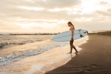 Okyanusta gün batımında beyaz sörf tahtası olan çekici genç bir sörfçü kadın. Bali Endonezya.