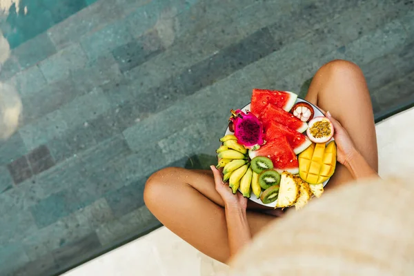 Joven mujer irreconocible junto a la piscina con un plato de frutas tropicales: sandía, piña, plátanos, mangostán, maracuyá, mango y fruta del dragón. —  Fotos de Stock