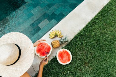 Havuzdaki genç çekici kadın olgun tropikal meyvelerin tadını çıkarıyor: karpuz, muz ve ananas..