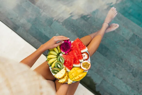 Fiatal, felismerhetetlen nő a medence mellett egy tál trópusi gyümölcsökkel: görögdinnye, ananász, banán, mangosztán, golgotavirág gyümölcs, mangó és sárkánygyümölcs. — Stock Fotó