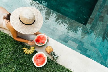 Havuzdaki genç çekici kadın olgun tropikal meyvelerin tadını çıkarıyor: karpuz, muz ve ananas..