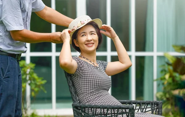 Asian middle aged couple sitting relax in backyard and putting on a hat to his wife — Stock Photo, Image