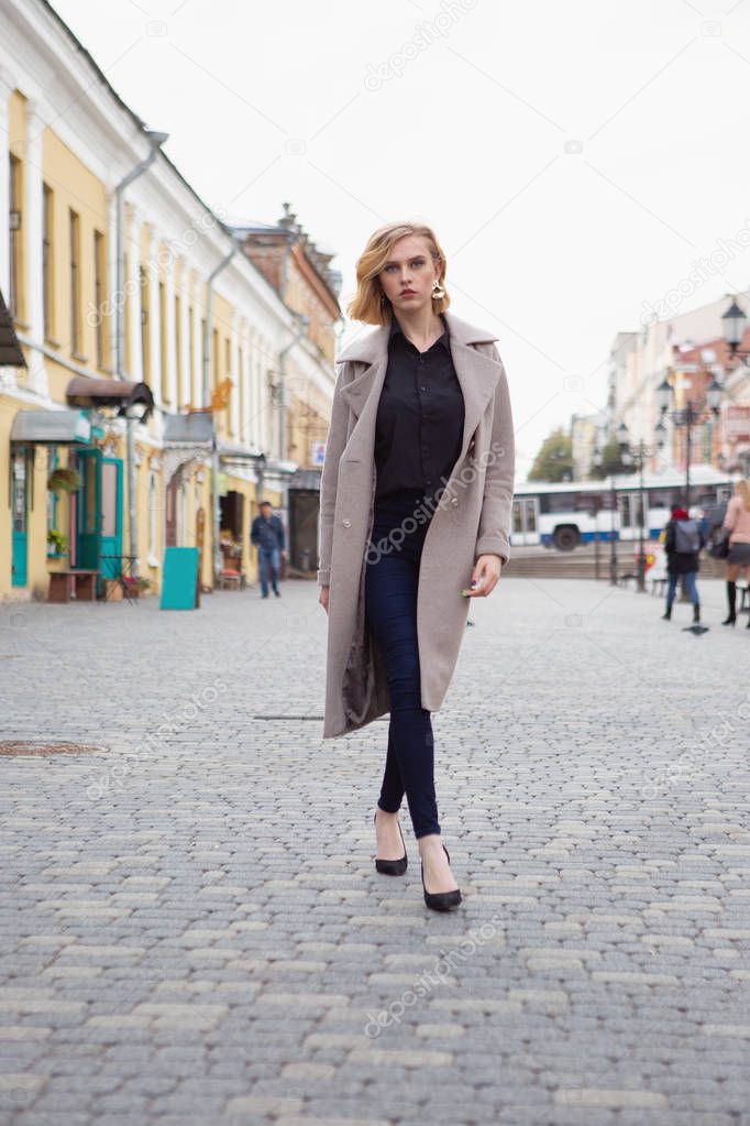 A charming blonde girl in an autumn coat walks around the autumn city