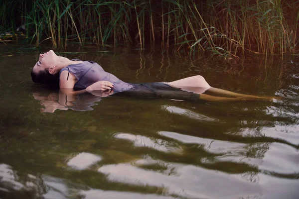 Koyu renk saçlı, ıslak gömlekli bir kız kafası geriye atılmış bir şekilde nehirde yatıyor.