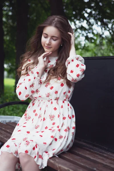 Uma mulher bonita em um vestido branco senta-se em um banco de parque em um dia ensolarado — Fotografia de Stock