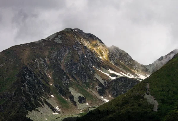 A mountain peak is lit by the sun rays coming through the cloudy dramatic sky.