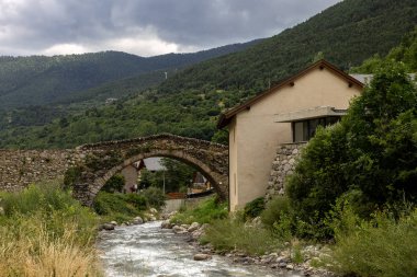 Eski bir taş köprü ön planda bir ev ile bir nehir geçer.