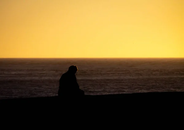 A seated silhouetted man looks down in front of the ocean before sunset.