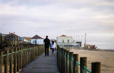 Plaja yakın bir tahta üzerinde bir orta yaşlı çift yürür. Bulutlu gün, arkadan görünüm.