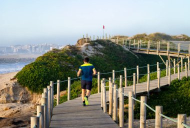 Genç adam sahil kum ve okyanusun bir kırmızı bayrak doğru çalışır. Mavi gömlekli. Arkadan görünüm. Açık gün.