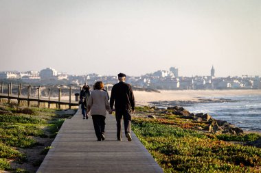 Üst düzey çift Okyanusu yakınlarındaki sahil boyunca yürüyerek. Uzak insanlar. Cityscape arka plan. Arka görüş. Kopya alanı.