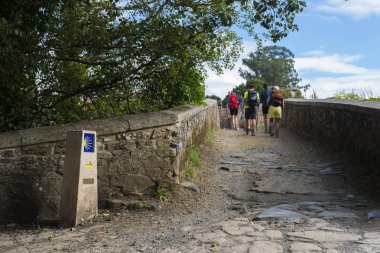 Furelos, İspanya - 31 Temmuz 2016: Sırt çantaları olan bazı genç hacılar bir ortaçağ köprüsünü geçerek Camino de Santiago'yu geçtiler..