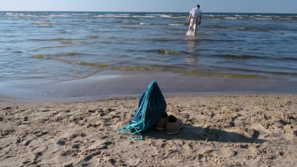 Man op ondiep water weglopen, rugzak en laarzen van links op het strand — Stockvideo