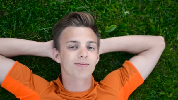 Teenager laying on the grass, smiles, winks to camera, arms behind head — Stock Video