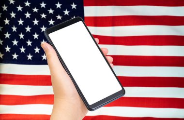 Hand 'in arka planında Amerikan ya da Amerikan bayrağı olan boş beyaz ekranlı akıllı telefonu tutarken yakın plan. Modifiye et ve uzayı kopyala