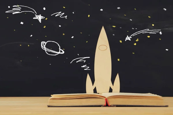 Back to school concept. wooden rocket and space sketchs over open book in front of classroom blackboard