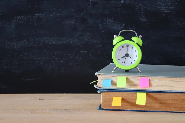 back to school concept. stack of books and alarm clock over wooden desk in front of blackboard