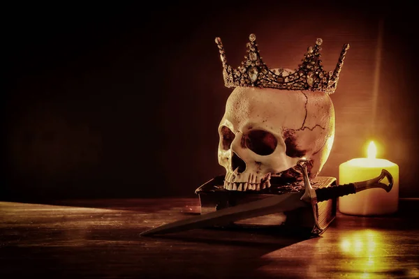 Calavera humana, libro viejo, espada, corona y vela ardiente sobre mesa de madera vieja y fondo oscuro . — Foto de Stock