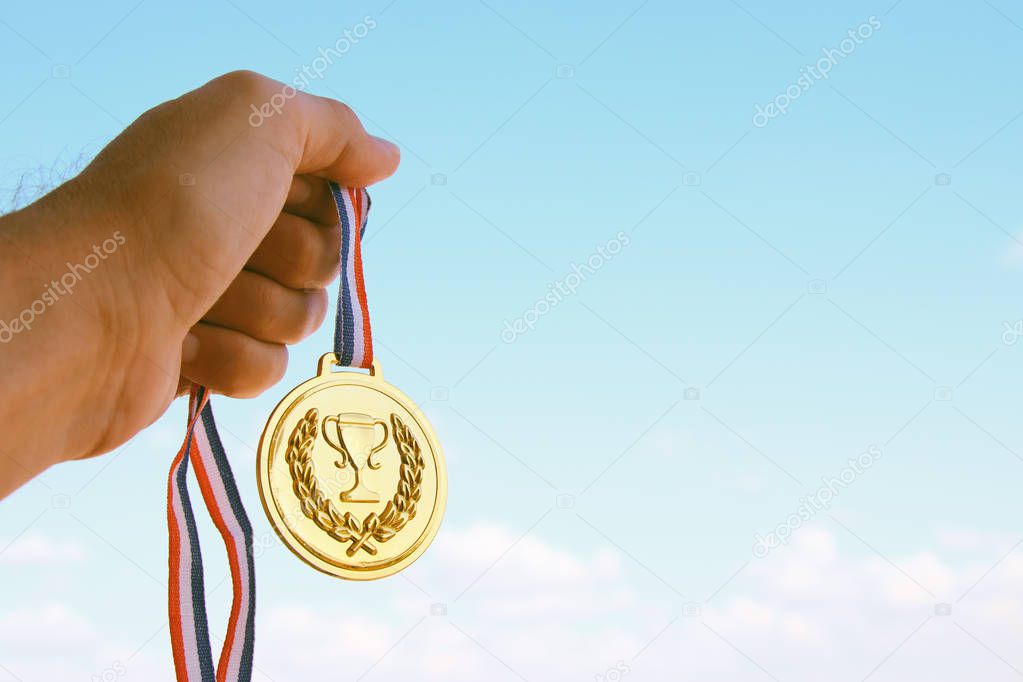 man hand raised, holding gold medal against sky. award and victory concept