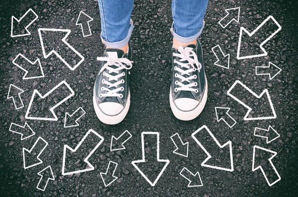 top view image of person in jeans and retro shoes standing over asphalt road with painted arrows showing different directions