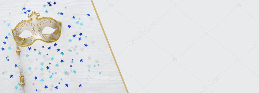 carnival party celebration concept with elegant gold mask on stick over white wooden background and stars. Top view