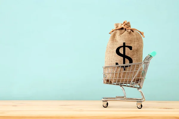 青い木製の背景上 袋いっぱいお金ドル記号とショッピングカート — ストック写真