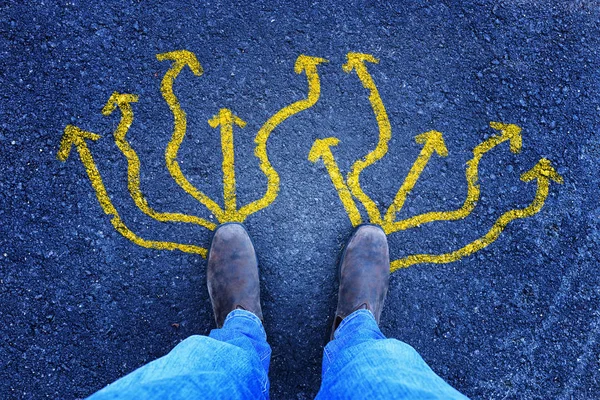 Foto vista superior de la persona en jeans y zapatos retro de pie sobre el camino de asfalto con flechas pintadas que muestran diferentes direcciones. Filtrado . —  Fotos de Stock