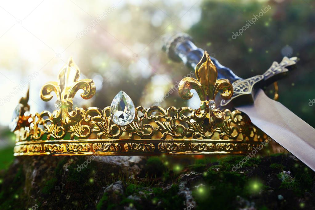 mysterious and magical photo of silver king crown and sword over the stone covered with moss in the England woods or field landscape with light flare. Medieval period concept