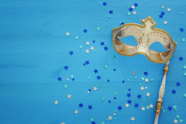 carnival party celebration concept with elegant gold mask on stick over blue wooden background and stars. Top view