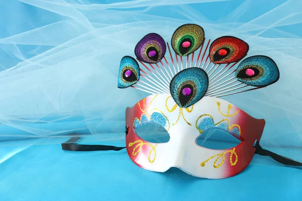 Photo of elegant and delicate venetian mask with peacock tail decoration element over turquoise silk background
