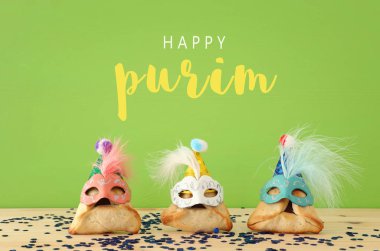 Purim kutlama kavramı (Yahudi karnaval tatil). Sevimli palyaço şapkalar ve ahşap masa ve yeşil arka plan üzerinde maskeleri ile geleneksel hamantaschen tatlılar