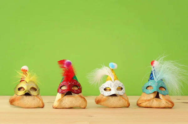 Purim viering concept (Joods carnaval vakantie). Traditionele hamantaschen cookies met leuke maskers over houten tafel en groene achtergrond. — Stockfoto