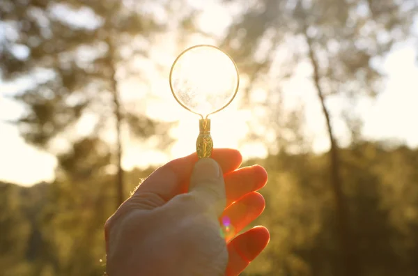 Una mano maschile tiene una lente d'ingrandimento contro il sole che tramonta all'esterno. concetto di ricerca, pensiero creativo e uso dell'energia solare — Foto Stock