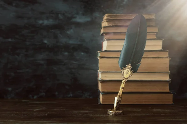 Old feather quill ink pen with inkwell and old books over wooden desk infront of black wall background. Vintage old style and filtered. Conceptual photo on history, fantasy, education and literature t — Stock Photo, Image