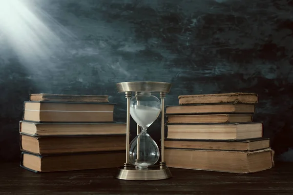 Hourglass as time passing concept and old books in front of black wall background. Conceptual photo on history, fantasy and education