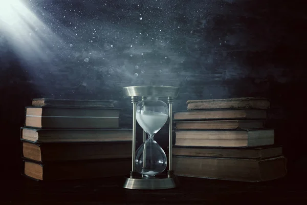 Reloj de arena como concepto de paso del tiempo y libros antiguos frente al fondo negro de la pared. Foto conceptual sobre historia, fantasía y educación —  Fotos de Stock