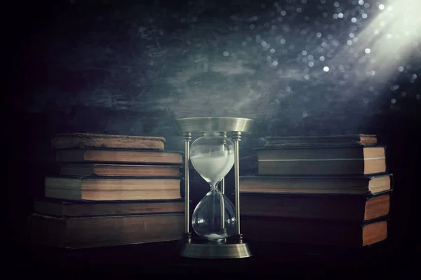 Reloj de arena como concepto de paso del tiempo y libros antiguos frente al fondo negro de la pared. Foto conceptual sobre historia, fantasía y educación —  Fotos de Stock