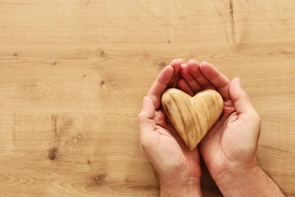 Ενήλικα αρσενικά χέρια που κατέχουν ξύλινη σχήμα καρδιά πάνω από το τραπέζι — Φωτογραφία Αρχείου