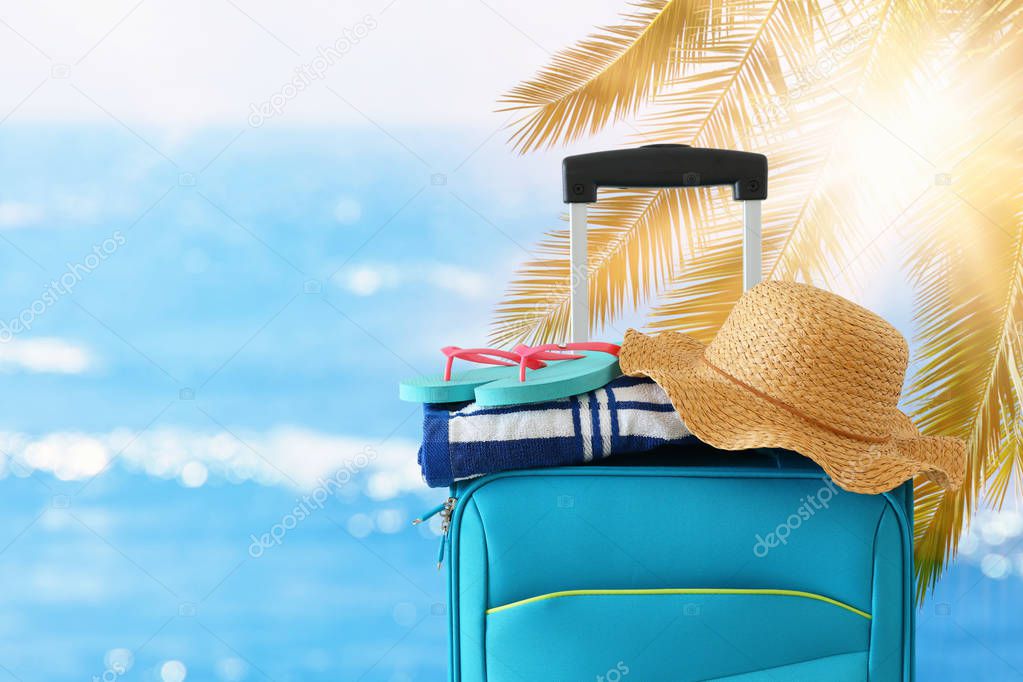holidays. travel concept. blue suitcase with female hat, flip flips and beach towel in front of tropical background