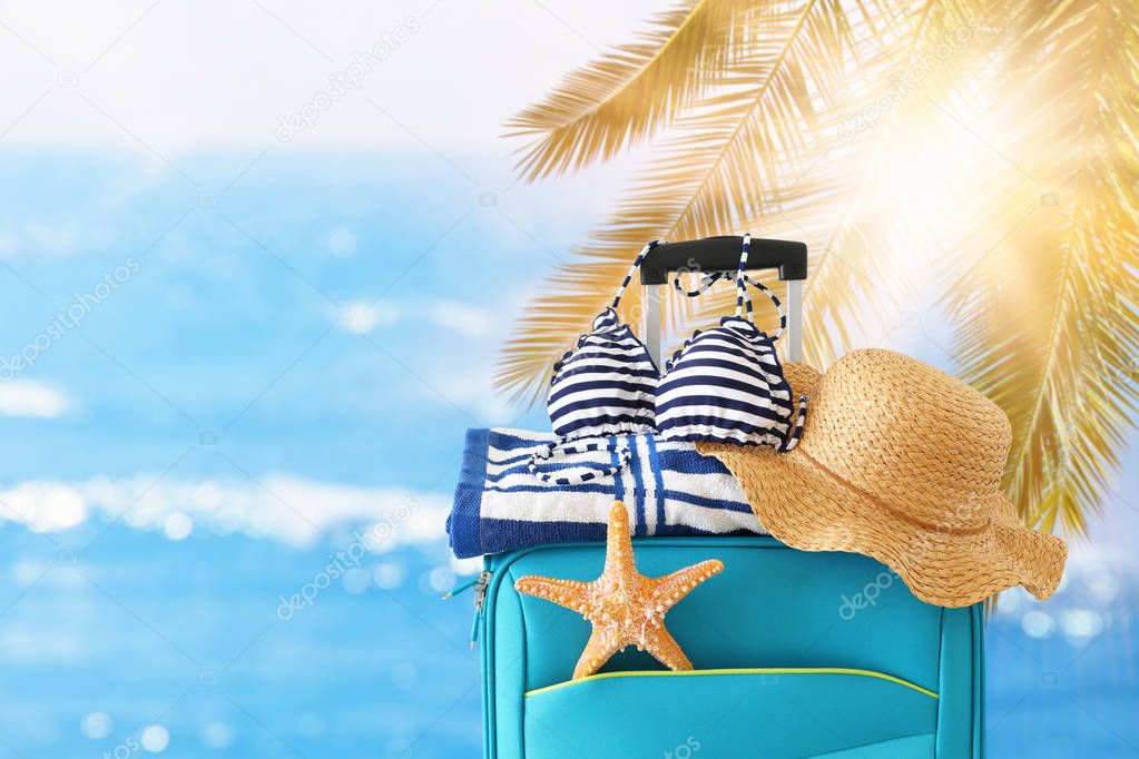 holidays. travel concept. blue suitcase with female hat, starfish, bikini and beach towel in front of tropical background
