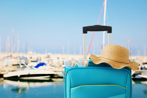 праздники. концепция путешествия. голубой чемодан с женской шляпой, солнцезащитные очки на фоне тропической марины
